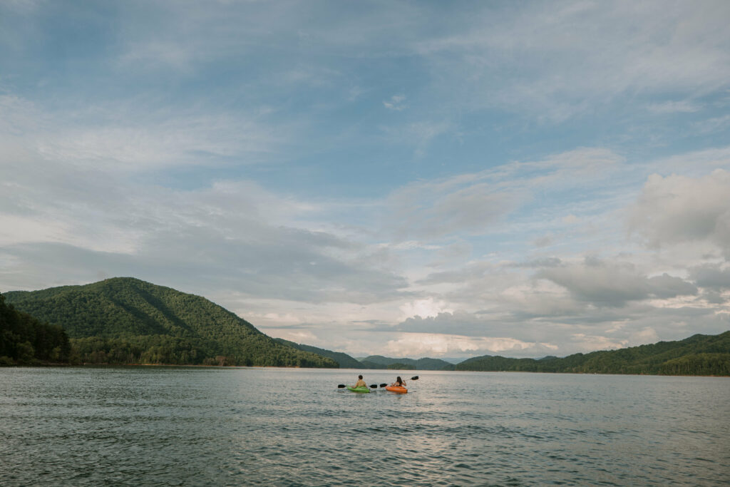 A couple is in the distance of a landscape mountain lake scene on their kayaks during their elopement day. The sky is full of clouds and they are in one green and one orange kayak. These eloping ideas include lots of water sports.