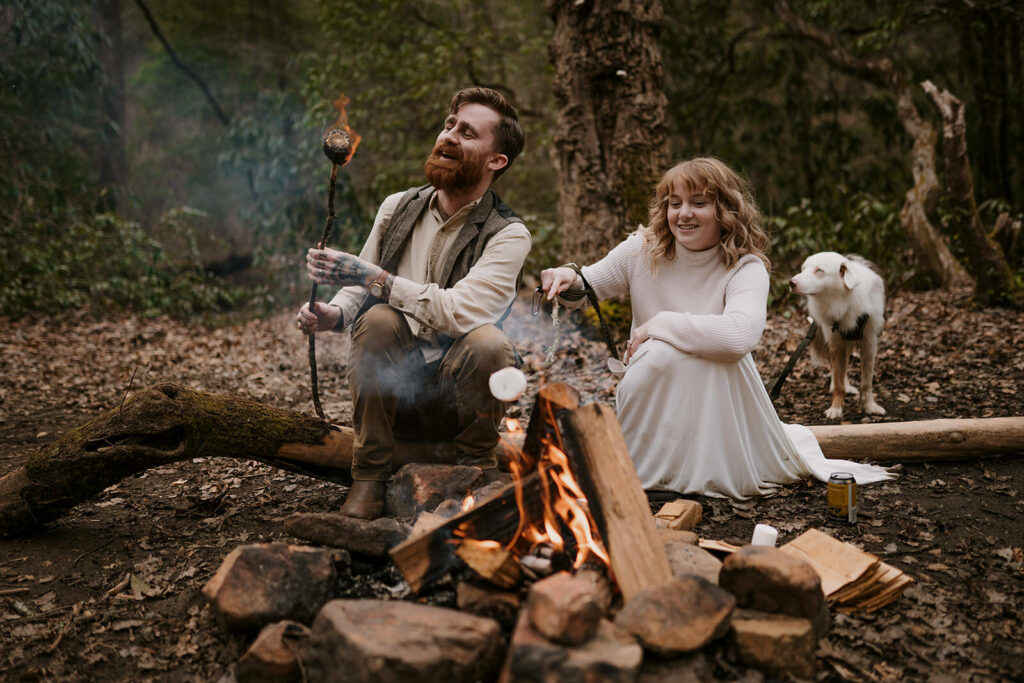 A couple sits on a log in front of a campfire in the evening. They are in casual elopement clothing and their dog is beside them. They are making s’mores over the fire and giggling. The grooms marshmallow is on fire and he is about to blow it out. Eloping ideas for outdoorsy couples can include simple activities like this too!