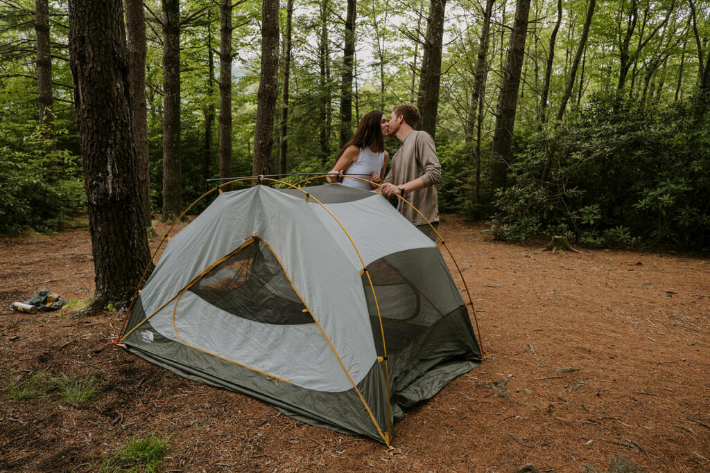 A couple sets up a tent in the woods on their elopement day. They are kissing behind the tent and the ground is covered in golden pine needles.
