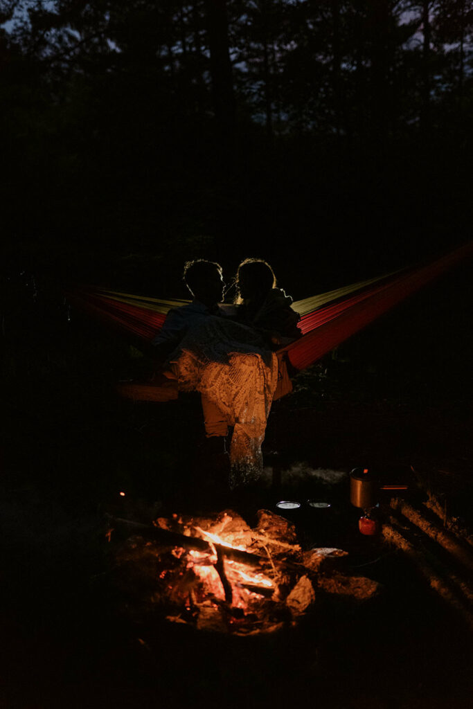 A couple sits in front of a campfire in their hammock at the end of their elopement day. They are silhouetted by light behind them.