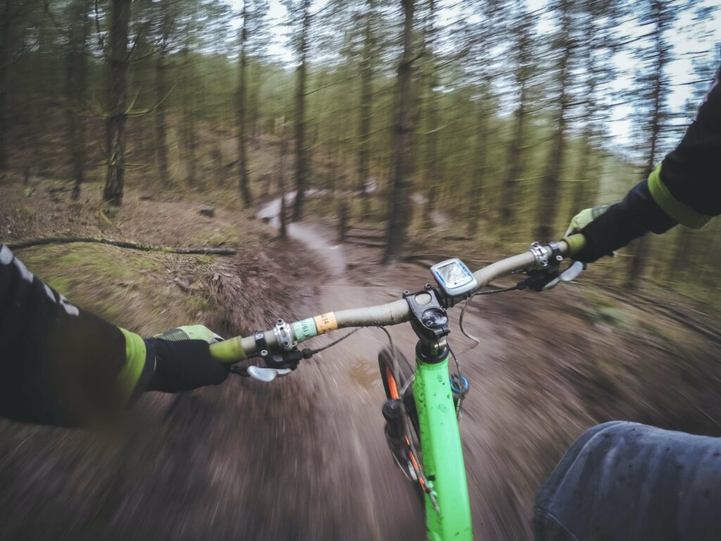A mountain biker is speeding down a trail in the forest. There is a view of their bars and hands. Mountain biking is a unique eloping idea.