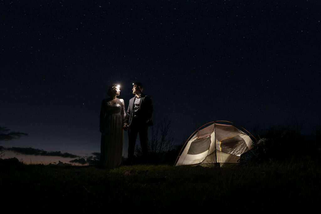 A couple stands beside their lit up tent looking at each other with headlamps on. They are underneath a starry sky and the sunset is behind them in the distance. They are wearing elopement wedding clothing because they just got married on the mountain. Eloping ideas for outdoorsy couples don't require a ton of movement.