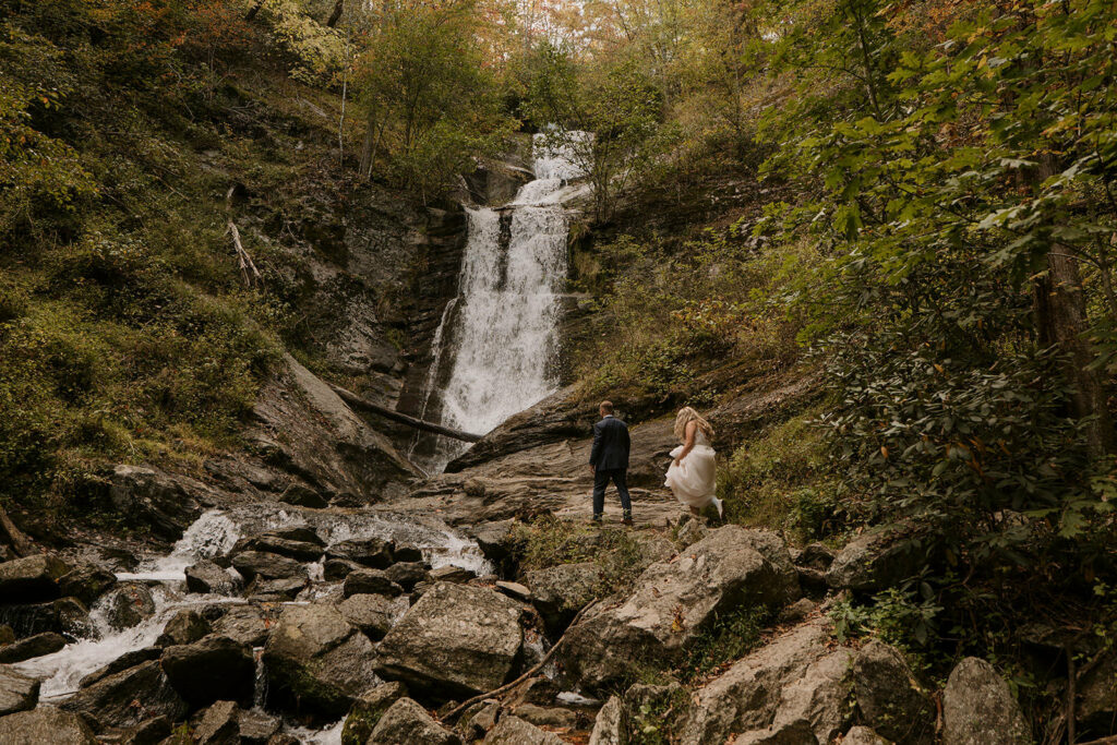 A couple walks up to the bottom of a waterfall on their elopement day in their dress and suit. The leaves are green all around them and the waterfall is tall and skinny.