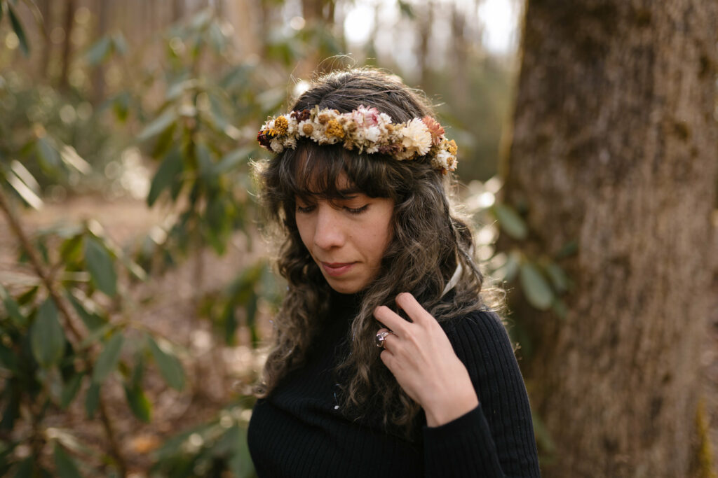 A bride is fixing her hair and adjusting her dried flower crown in the woods. She is wearing a black long sleeve shirt and standing in front of a tree and rhododendrons.