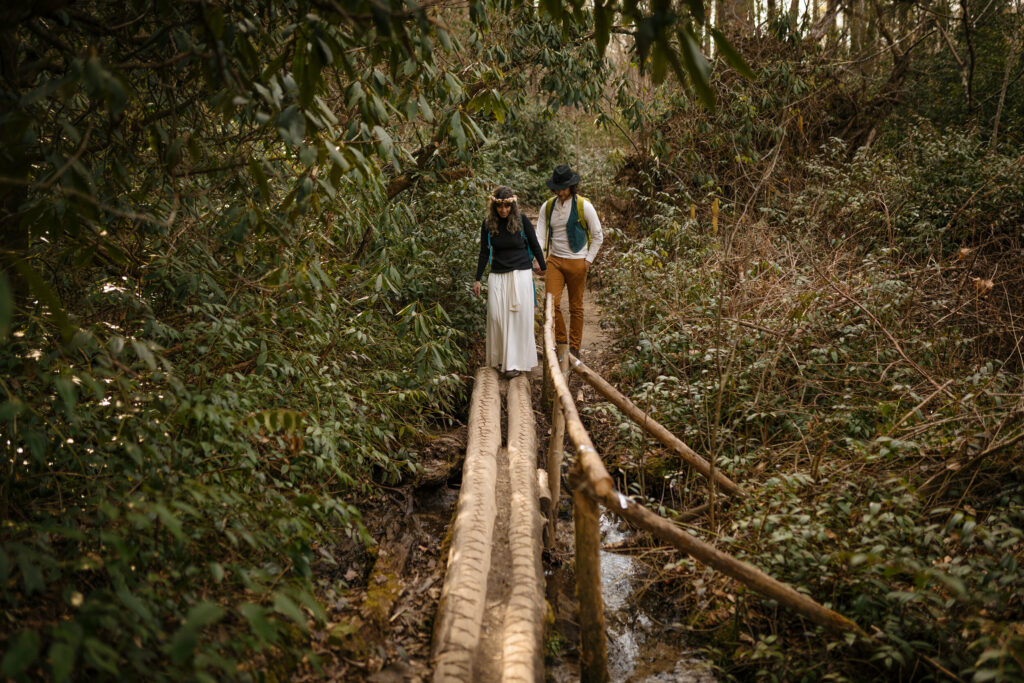 A couple in their elopement clothes and hiking backpacks is crossing a natural bridge above a creek in the forest.