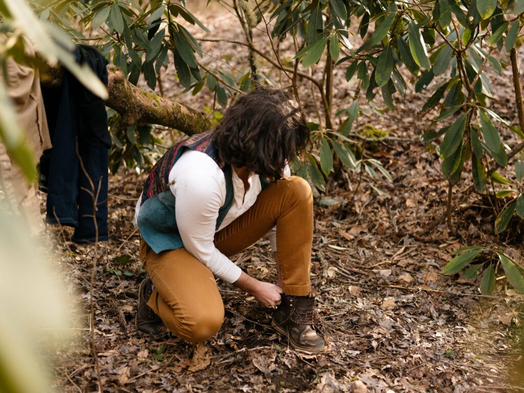 A guy crouches down and puts his boots on in the forest surrounded by rhododendron bushes. His previous clothes are hanging on a nearby branch.