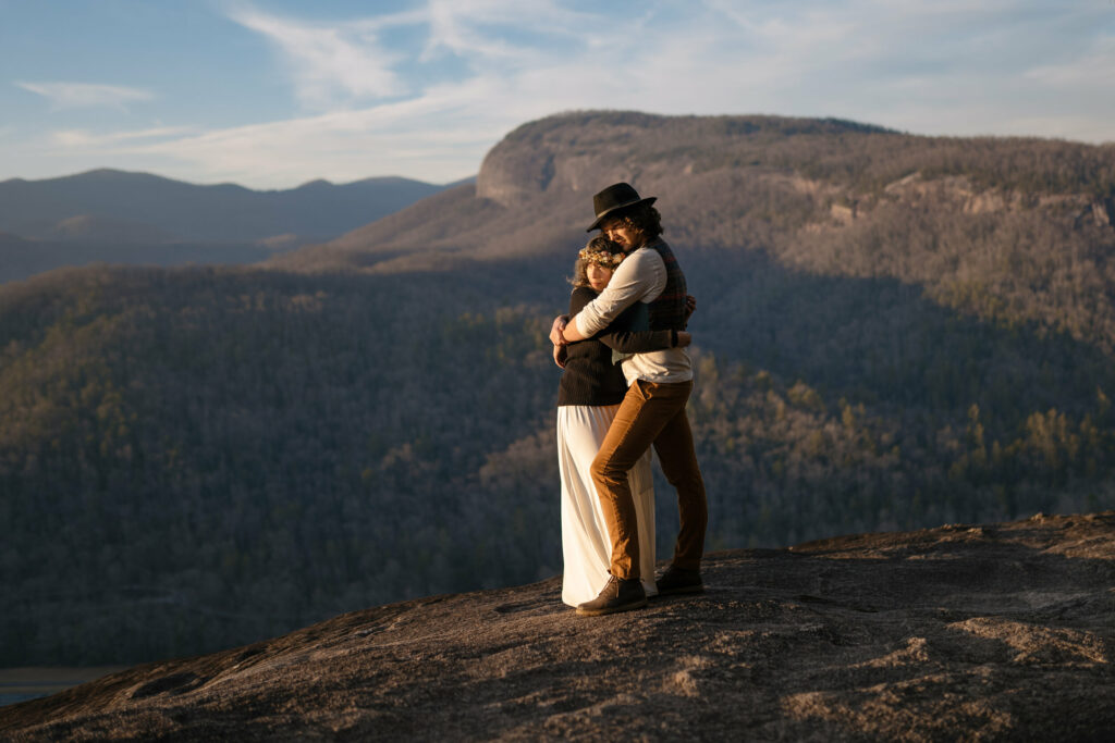 A couple is standing in front of a big rock outcropping on a rock face at sunset hugging. The girl is wearing a flower crown and the guy is wearing a black hat.