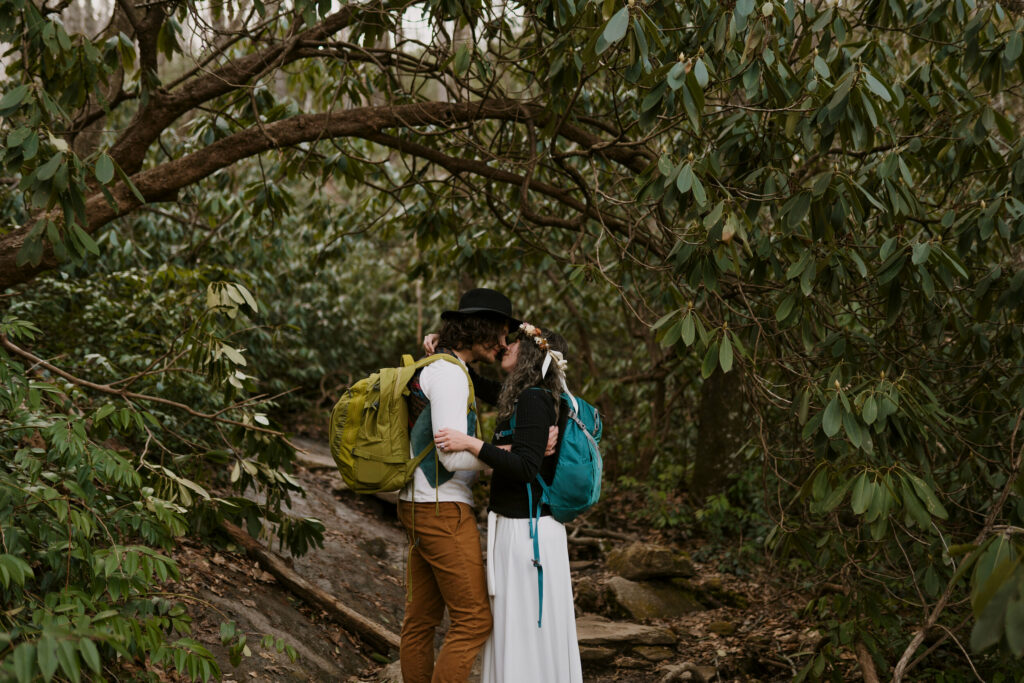 During their hiking elopement, a couple stops beneath the rhododendron branches to hug and connect with their hiking backpacks on.