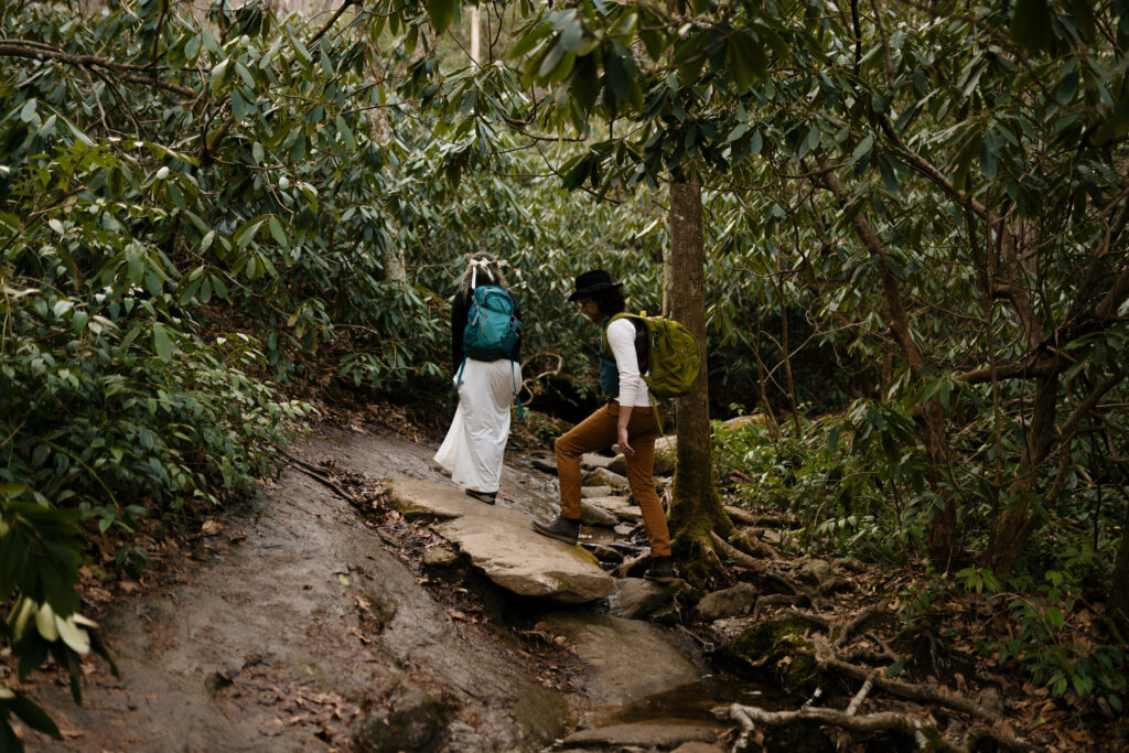 A couple is hiking with their backpacks on and elopement clothes through rhododendron tunnel, and over rocks and roots.