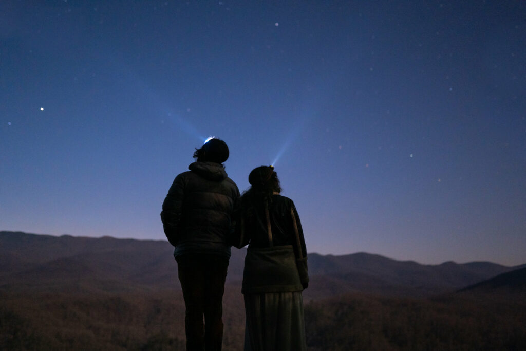 A couple stands with their headlamps toward the sky looking out at the starry night time mountain landscape.