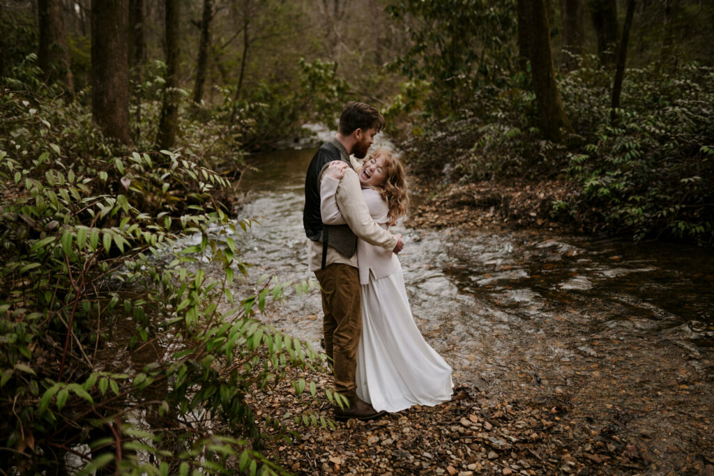 A couple is hugging and laughing during their elopement beside a river in the forest. If you want to know how to plan your elopement you can do so in the winter with simple clothes on like them!