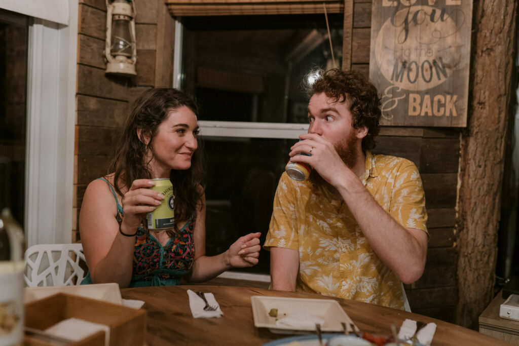 A couple looks at each other with goofy faces enjoying a beer at the end of their elopement day.