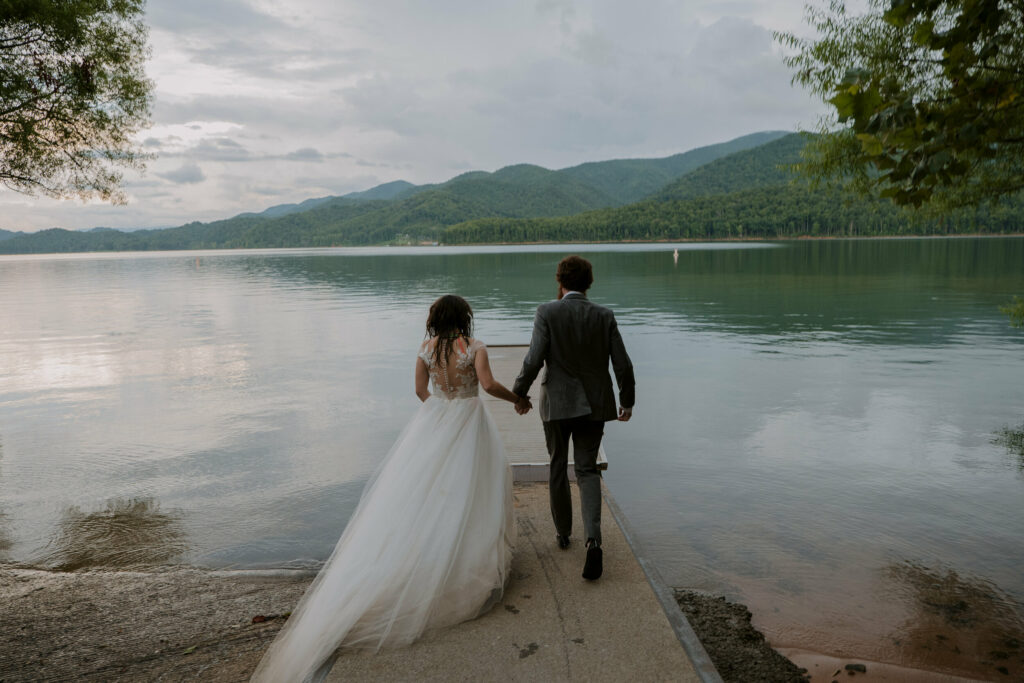 A couple is running towards a boat dock in their wedding clothes surrounded by mountains at sundown.