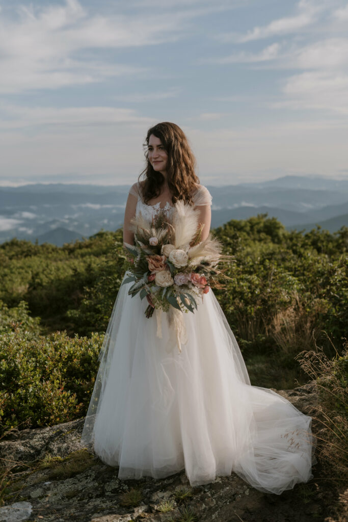 A bride stands in front of a mountain scene with her silk green, pink, and beige bouquet for her elopement day.