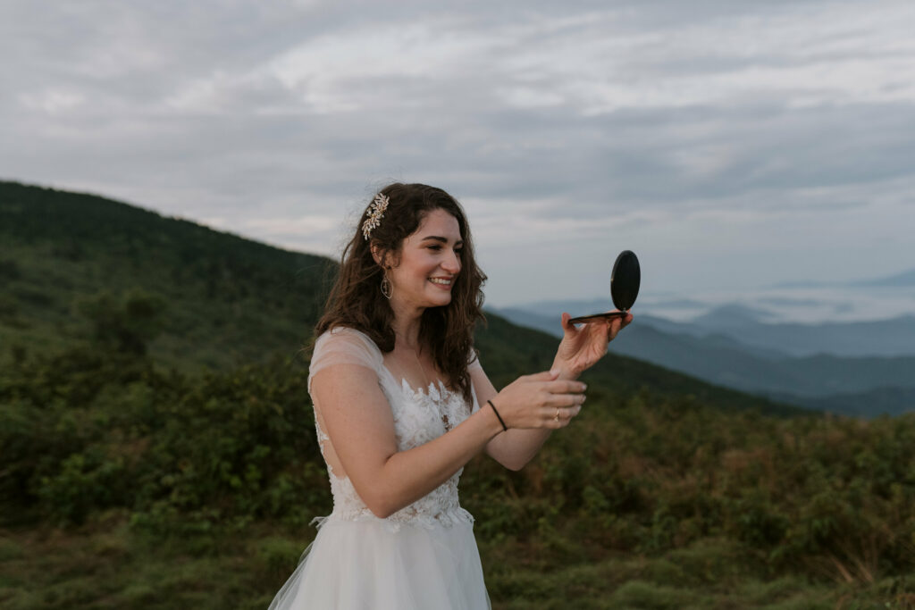 A bride is getting ready in her wedding dress on top of a mountain for her elopement. She is looking into a compact mirror and smiling. The sun hasn’t risen and the light is blue and soft.