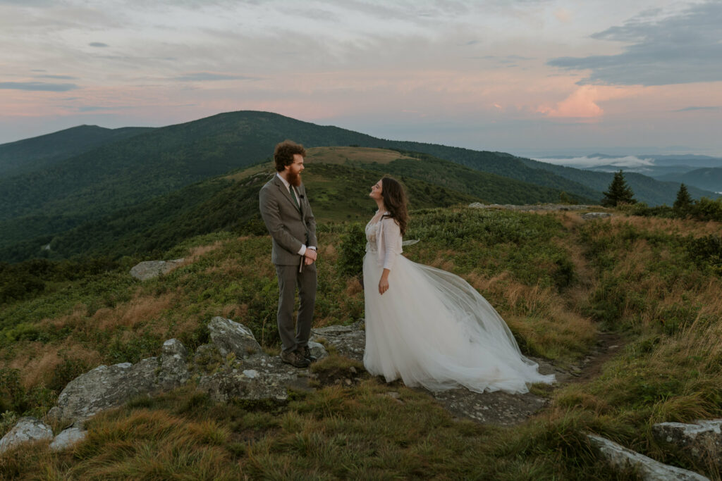 A couple stands on a mountaintop in the blue ridge mountains for their elopement. They are facing each other about to read their vows at sunrise.
