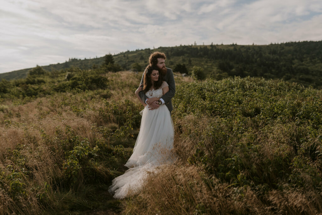 A groom hugs his bride from behind amidst a high grassy field on a mountain during the morning of their elopement.