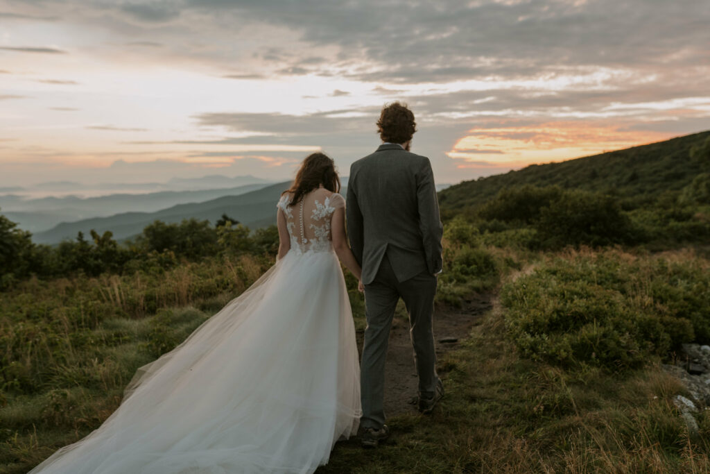 A couple walks toward the sunrise on a mountain, on in their elopement wedding dress and the other in a suit.
