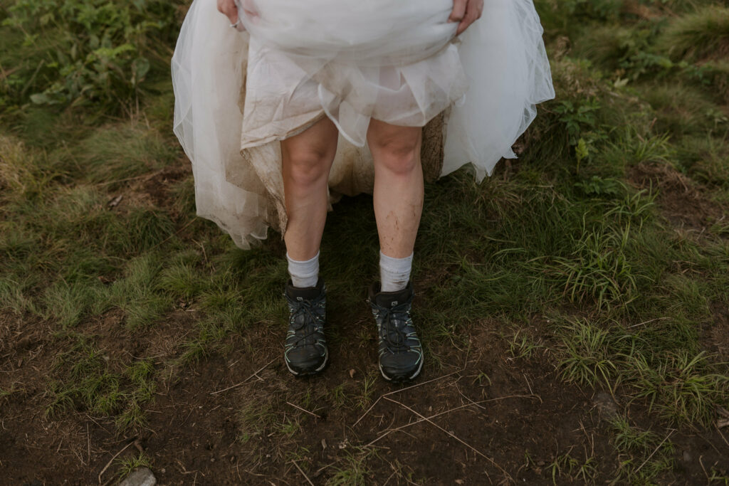 A bride holds her dress up to show off her muddy legs and hiking boots for her mountain elopement.