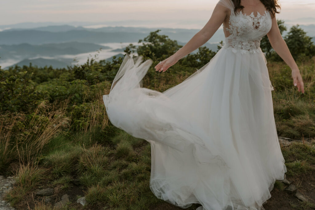 The bottom of a white elopement dress is being tossed out to the side by a bride in the grass on a mountaintop.