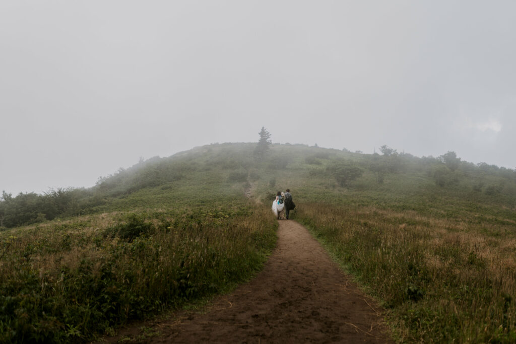A small bride and groom during their mountain elopement are hiking on a trail in the distance while fog moves over the mountain.