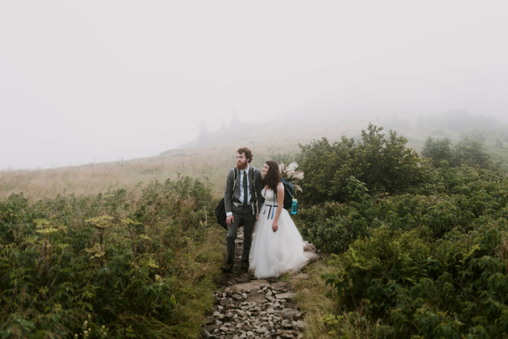 A couple stands on a trail surrounded by green bushes  facing a foggy mountain view during their elopement with their wedding clothes and backpacks on.