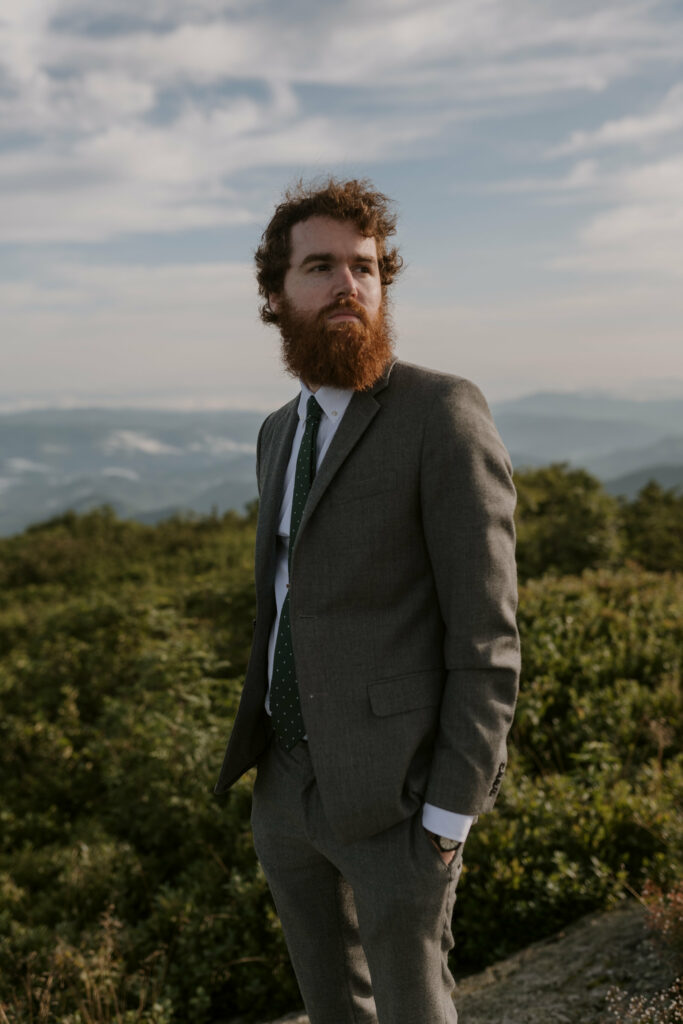 A groom poses in front of a mountain scene in a grey suit on his elopement day.