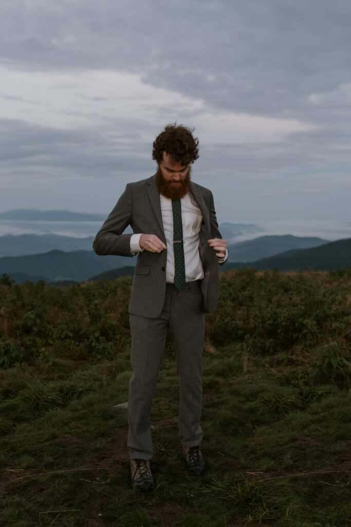 A groom is getting ready and buttoning his jacket on top of a mountain for his elopement. It is right before sunrise and the light is blue and soft.