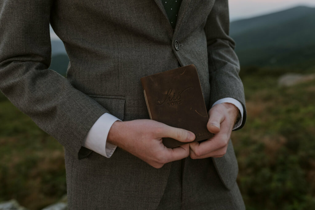 A groom holds his vow book in his hand on a mountain during his elopement.