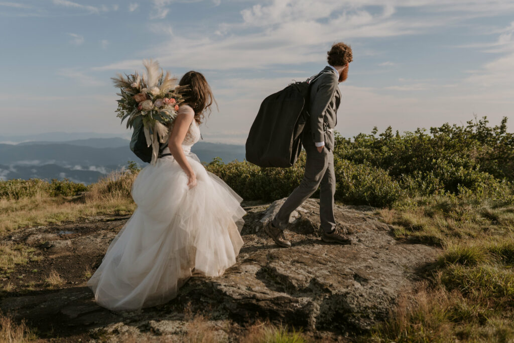 A couple in their elopement clothing and backpacks starts a hike on a mountaintop.