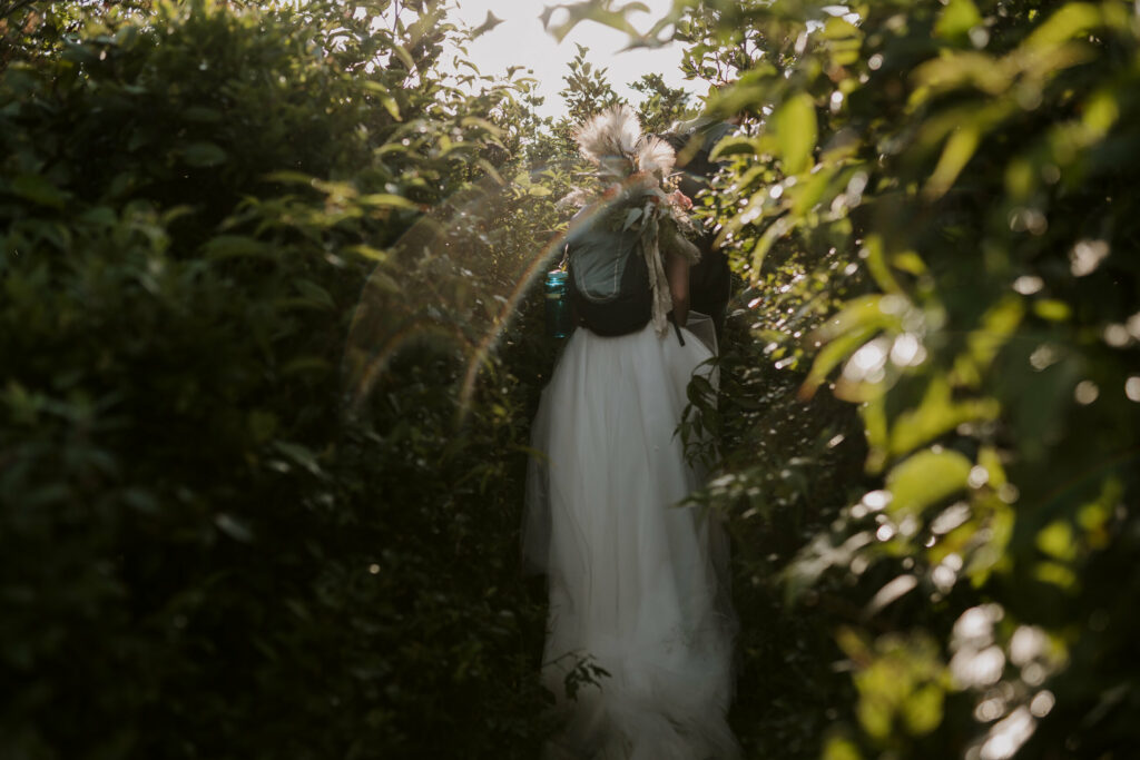 A bride walks through big bushes with her backpack on and her long white dress grazing the trail behind her.