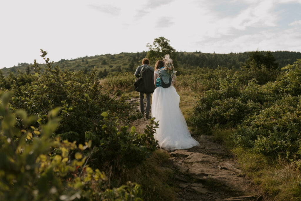 A bride and groom hike on a trail across a mountaintop in elopement clothing and backpacks on.