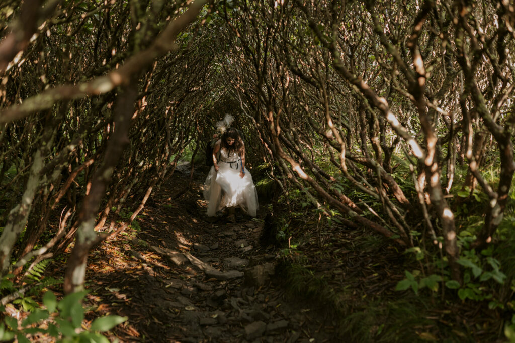 A bride is hiking in her elopement dress and backpack through a big tunnel of rhododendrons with craggy branches and light streaming through.