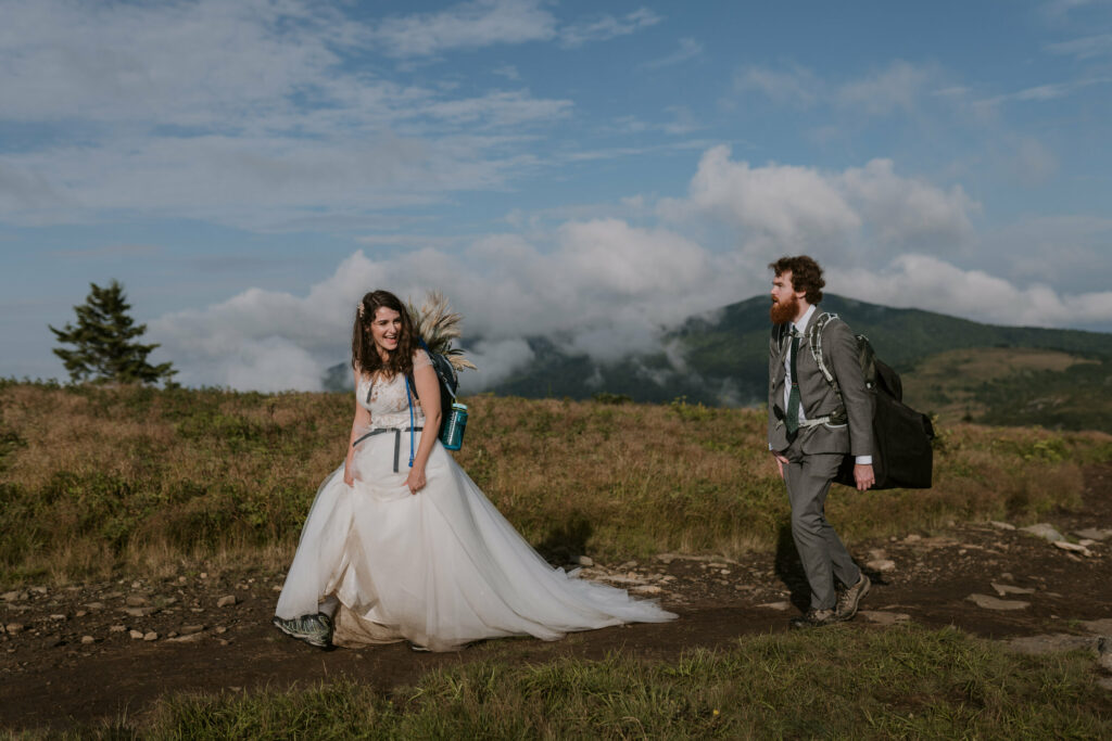 A bride and groom hike on a trail across a mountaintop in elopement clothing and backpacks on. The bride is looking back and smiling.