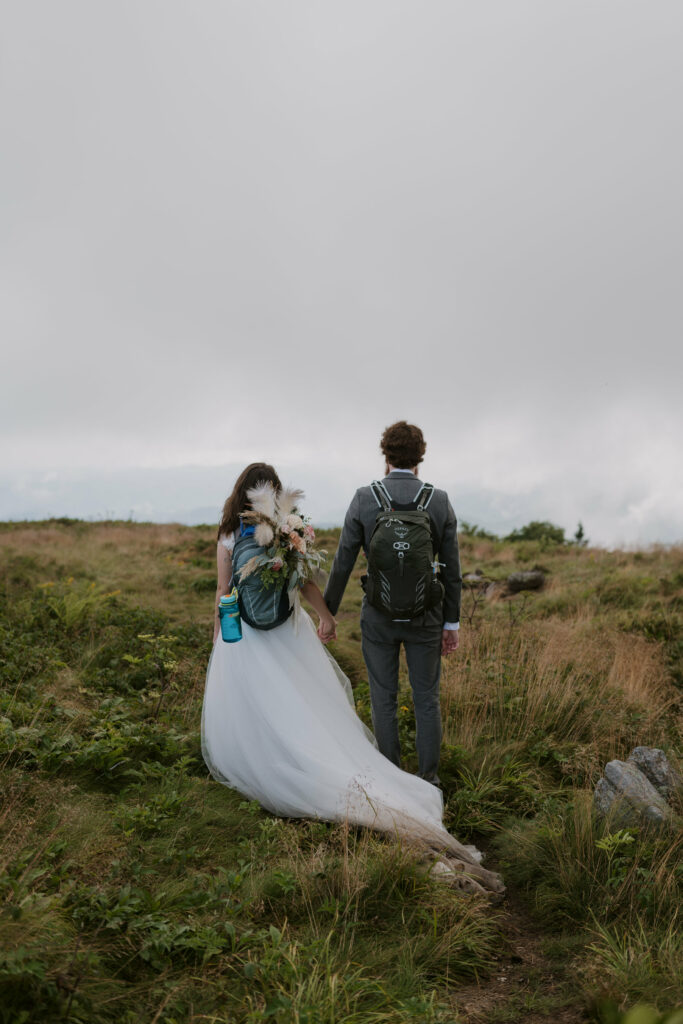 A couple stands facing a cloudy mountain view during their elopement with their wedding clothes and backpacks on.