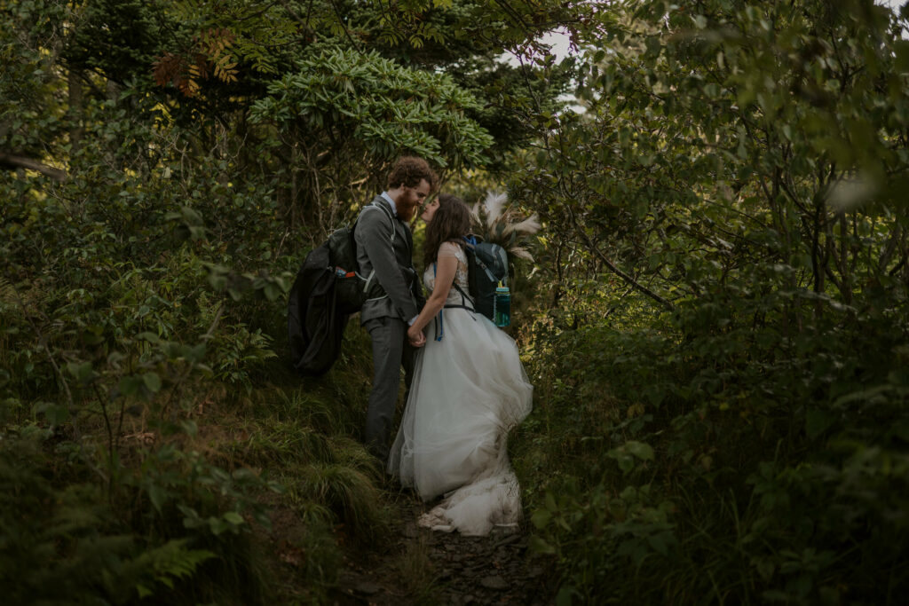 A bride and groom stand face to face holding hands in their wedding clothes and backpacks. They are in the middle of a trail surrounded by green trees smiling at each other.
