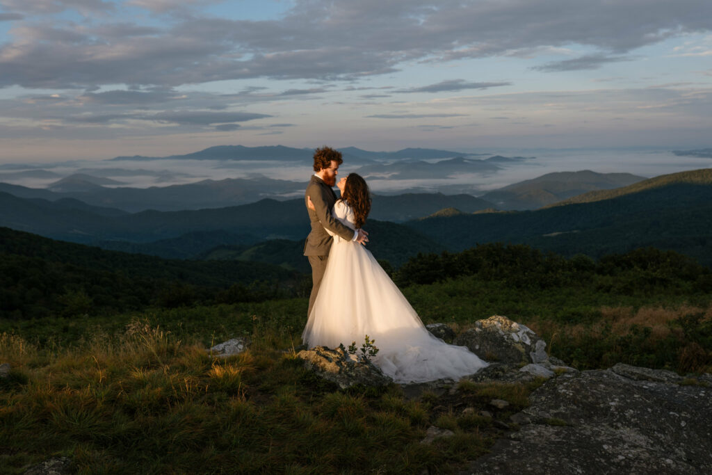 A couple stands in the sunshine on a mountain during their elopement holding each other and looking at each other. There is a beautiful blue and green mountain scene in the background and they are in wedding clothes.