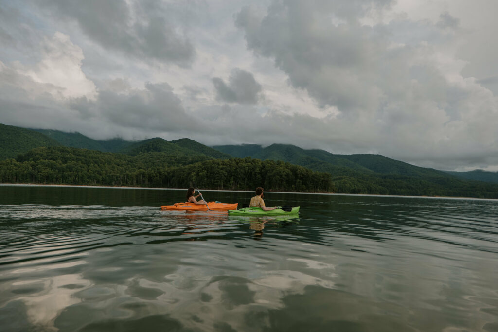 A couple sits in orange and green kayaks on a lake surrounded by mountains on a cloudy afternoon as a part of their elopement day.