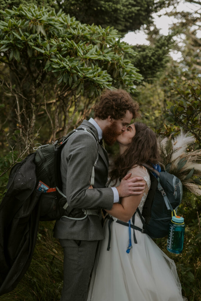 A bride and groom stand face to face holding hands in their wedding clothes and backpacks. They are in the middle of a trail surrounded by green rhododendron kissing.