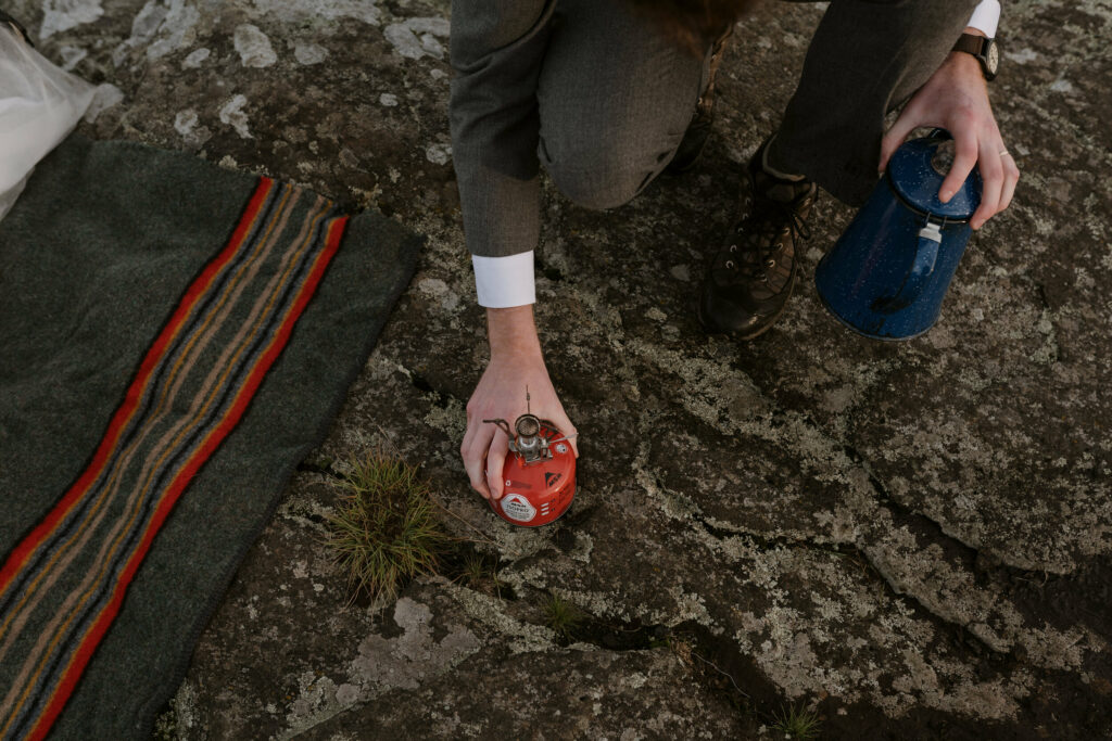 A close up of a groom holding a blue percolator and a red fuel canister crouch beside a striped blanket on a rock during his elopement day.