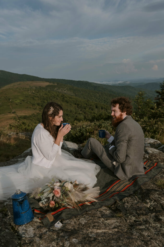 A bride and groom are sitting on a blanket on a mountain with their elopement clothes on smiling and sipping their coffee.