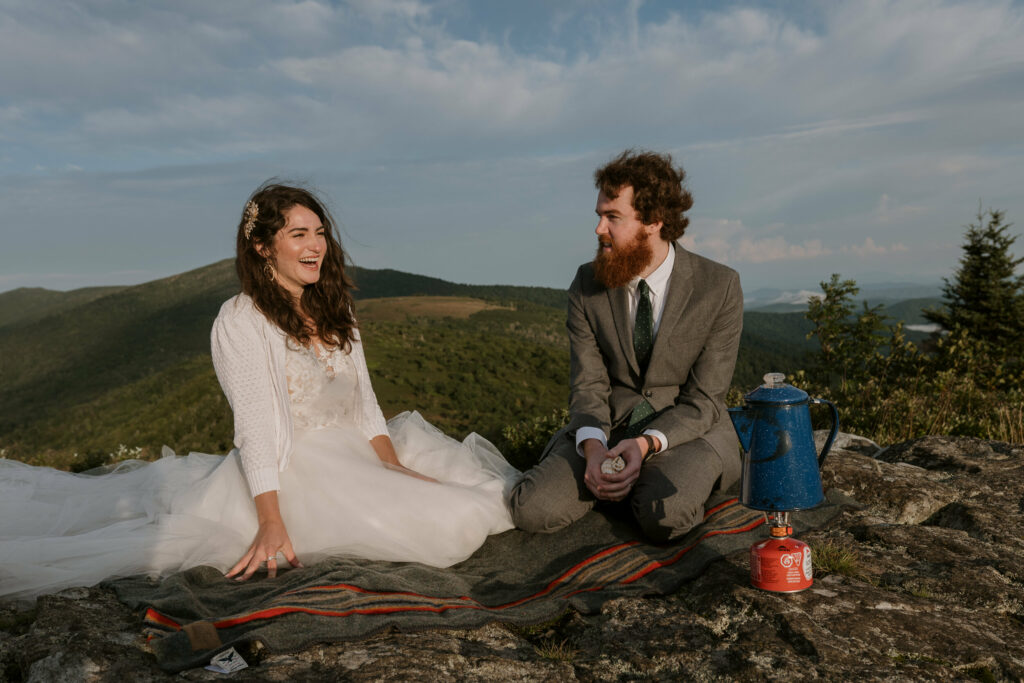 A bride and groom are sitting on a blanket on a mountain with their elopement clothes on laughing while making coffee.