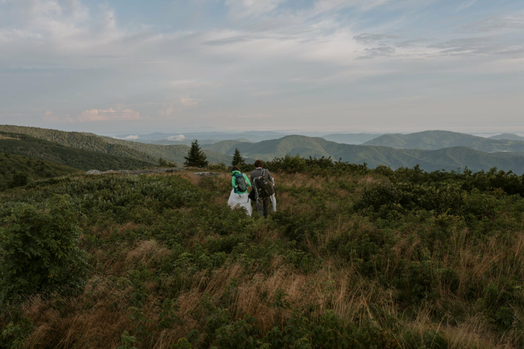 A couple wearing elopement clothes and backpacks walks through high grass on a mountaintop on a partly cloudy day.