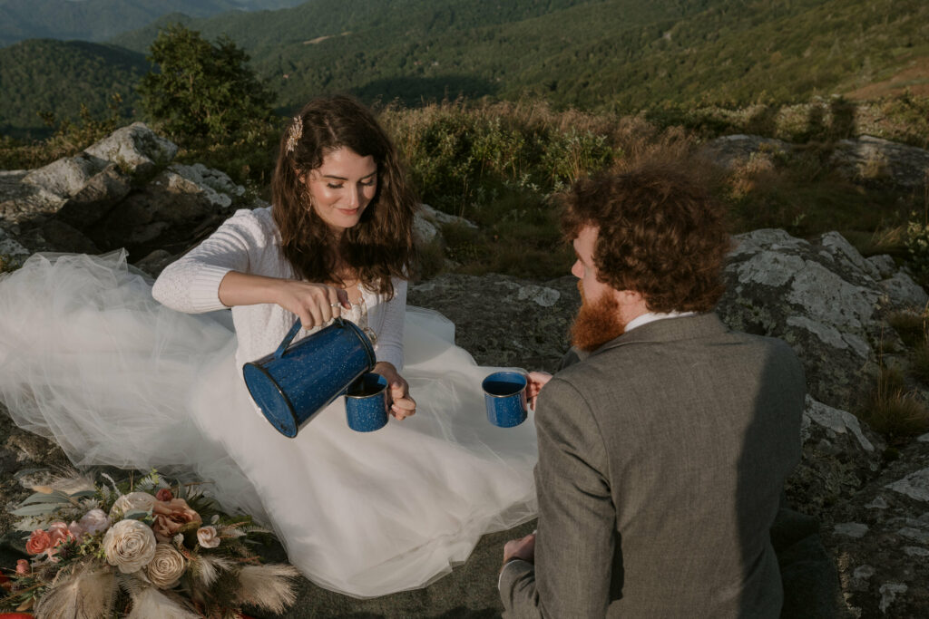 A bride pours out some coffee from a percolator into her mug while sitting on a mountain across from her groom holding another mug of coffee during their elopement day.