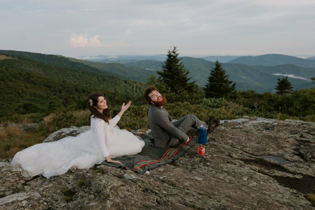 A bride and groom are sitting on a blanket on a mountain with their elopement clothes on laughing.