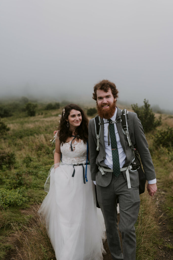 A bride and a groom walk toward the camera in their elopement clothing and backpacks on a mountain. The bride is smiling at the groom and the groom is smiling at the camera.
