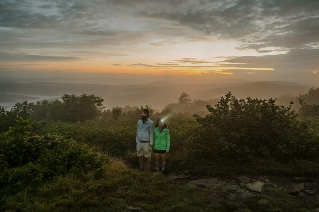A couple stands on mountain at the beginning of their elopement day, holding hands and wearing shining headlamps. The sun is rising over the mountains behind them with a blue, pink, and orange cloudy sky.