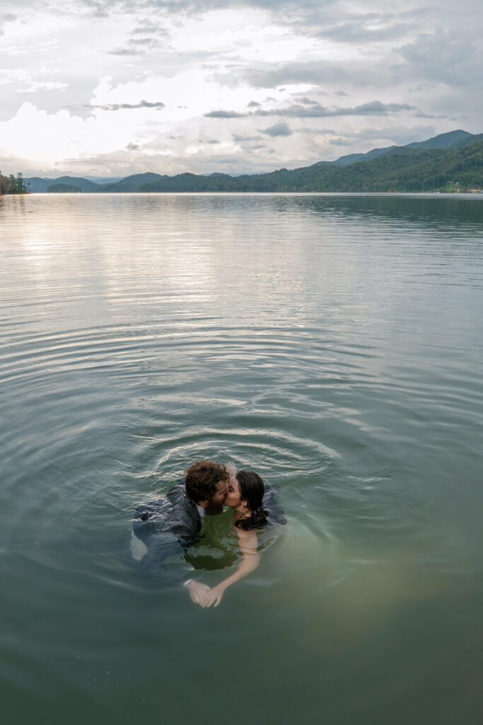A couple is swimming in their elopement clothing in a blue lake surrounded by mountains at sunset.