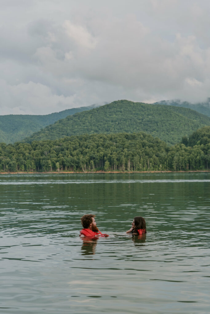 A couple swims in a lake with life jackets on surrounded by mountains on a cloudy afternoon as a part of their elopement day.