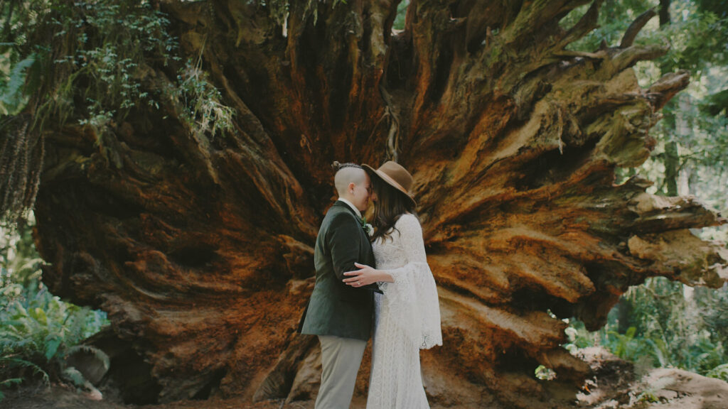 A couple stands in their elopement clothing in front of a massive downed redwood tree during their wedding day. They are forehead to forehead.
